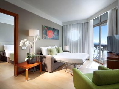 Jasny pokój hotelowy z łóżkiem dwuosobowym, zielonym fotelem, biurkiem i balkonem z widokiem na morze.