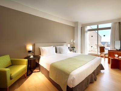 Jasny pokój hotelowy z dużym łóżkiem, zielonym fotelem i balkonem z widokiem na morze.