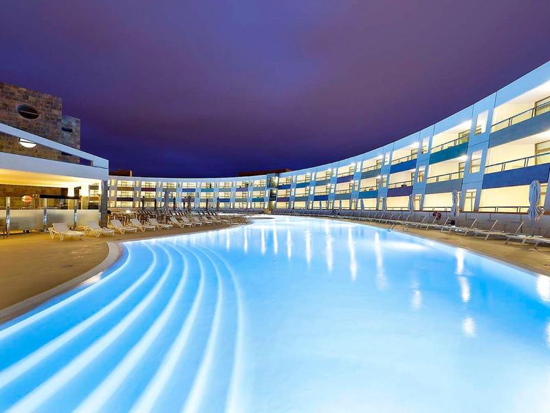 Nowoczesny hotel z dużym podświetlanym basenem na zewnątrz nocą.