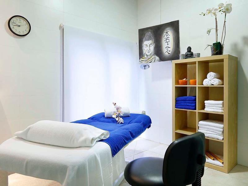 Nowoczesny pokój zabiegowy z łóżkiem do masażu, krzesłem i półką z ręcznikami i dekoracjami.