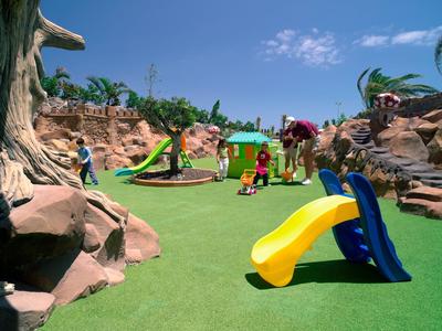 Bunter Spielplatz mit Rutschen und Sandbereich vor einem Hotel unter blauem Himmel.