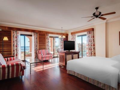 Helles Hotelzimmer mit Holzfußboden, Bett, Sofa, TV und großen Fenstern mit Blick auf Balkon.