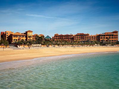 Strand mit klarem Wasser und großen Hotelgebäuden im Hintergrund unter blauem Himmel.