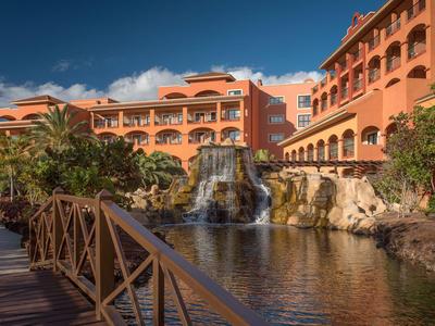 Hotel mit orangefarbenem Gebäude, Wasserfall und See unter blauem Himmel.