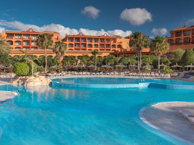 Großer Hotelpool mit klarem Wasser, umgeben von Palmen und sonnigen Hotelgebäuden.