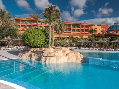 Großer blauer Pool vor einem Hotel mit Palmen und Sonnenschirmen unter blauem Himmel.