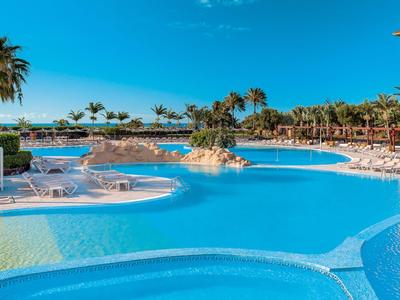 Großer Hotelpool mit Sonnenschirmen, Liegestühlen und Palmen unter klarem blauem Himmel.