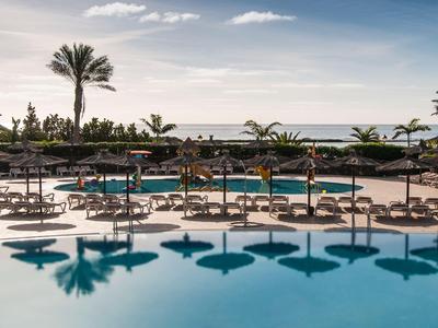 Hotelpool mit Sonnenschirmen und Palmen vor ruhigem Meer unter blauem Himmel.