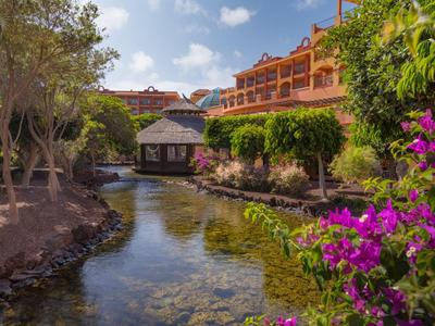 Hotelanlage mit Fluss, Bäumen, bunten Blumen und blauem Himmel.