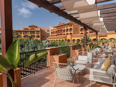 Hotelterrasse mit weißen Sitzmöbeln und Blick auf grünen Garten unter blauem Himmel.