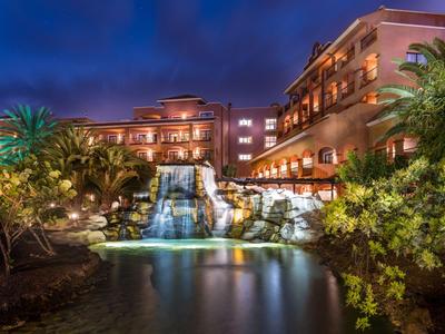 Beleuchtetes Hotel mit Wasserfall und Teich in tropischer Gartenanlage bei Nacht.