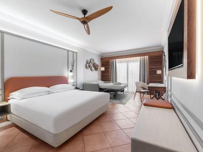 Helles Hotelzimmer mit großem Bett, Deckenventilator und Balkon mit Blick ins Freie.