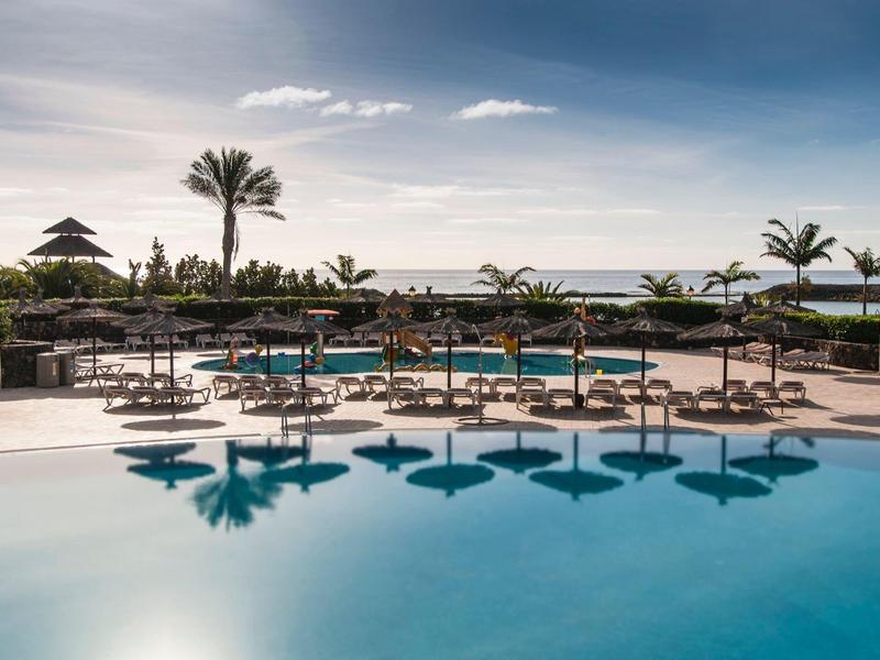 Groot zwembad met parasols en palmbomen onder een blauwe lucht in een resort.