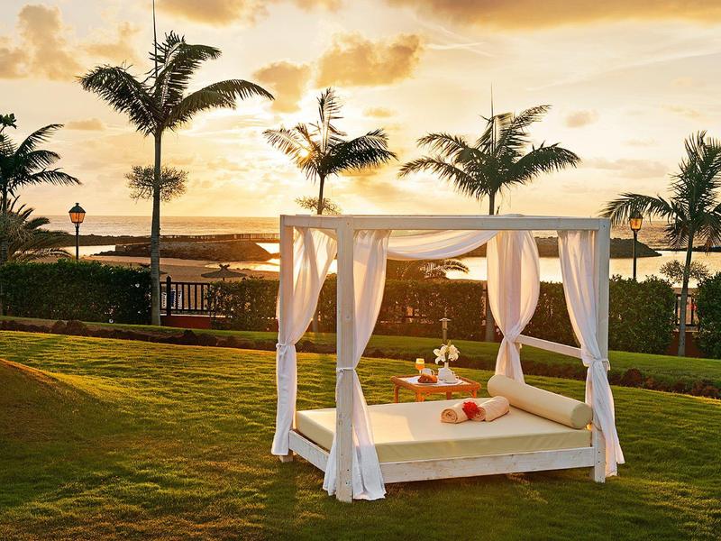 Een luxe wit hemelbed op een groen gazon bij zonsondergang met palmbomen op de achtergrond.