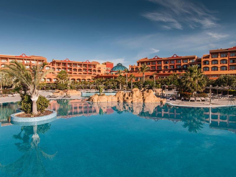 Groot hotelzwembad met palmbomen en parasols voor meerverdiepingen hotelgebouw onder blauwe lucht.