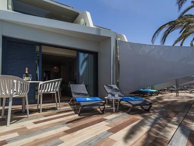 Terrasse mit blauen Liegestühlen, weißem Tisch und Stühlen vor modernem Haus bei Sonnenschein.