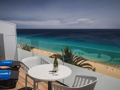 Balkon mit weißen Stühlen, Tisch und Weinflasche, Blick auf Strand und türkisfarbenes Meer unter bewölktem Himmel.
