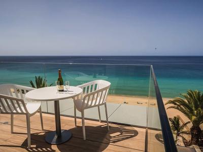 Terrasse mit weißem Tisch, zwei Stühlen, Weinflasche und Glas, Blick auf Meer und Sandstrand.