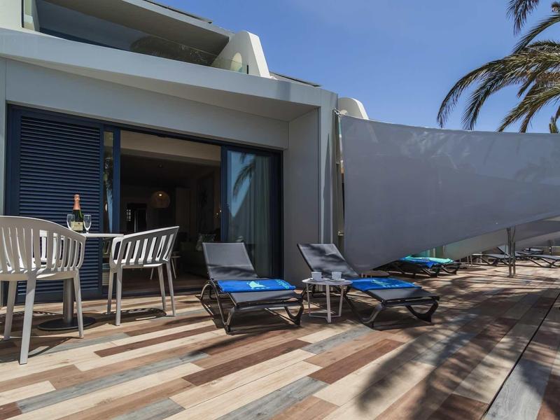Terrasse mit blauen Liegestühlen, weißem Tisch und Stühlen vor modernem Haus bei Sonnenschein.