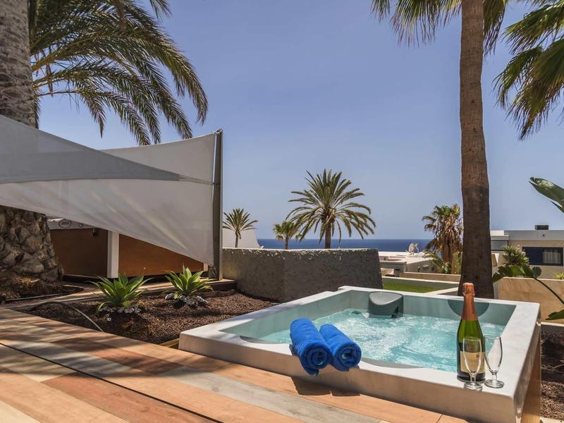 Kleiner Pool mit blauem Handtuch, Sektflasche und Palmen unter blauem Himmel am Meer.