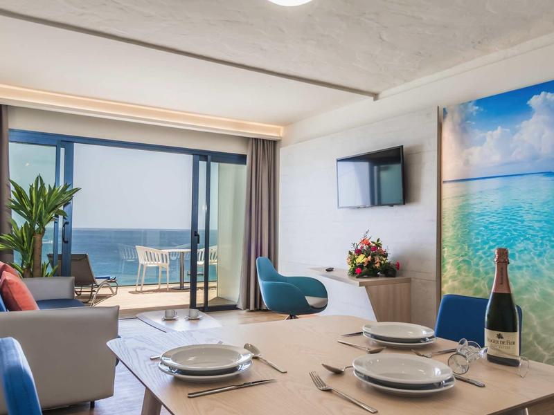 Essbereich mit gedecktem Tisch, blauen Stühlen, Pflanzen und Meerblick durch große Fenster.