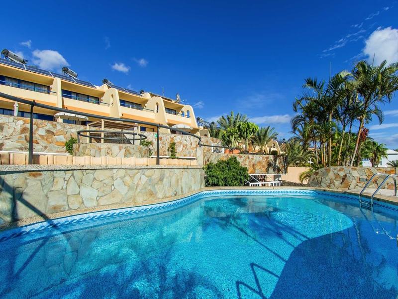 Großer blauer Pool vor mehrstöckigem Haus mit Terrasse, Palmen und klarem Himmel.