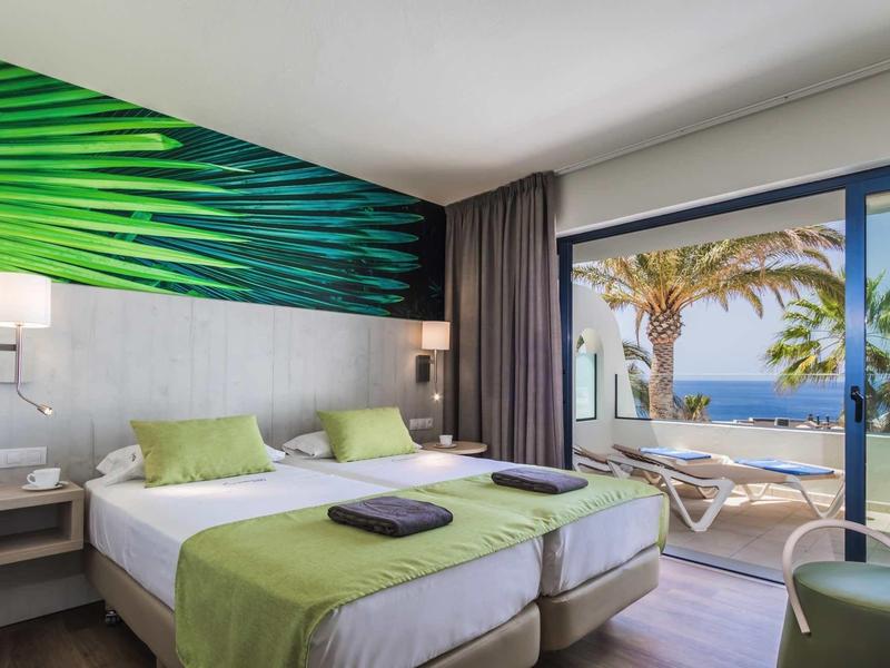 Helles Hotelzimmer mit zwei Betten, grünem Wandmotiv, Balkon und Blick auf Palmen und Meer.