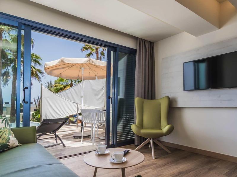 Modernes Wohnzimmer mit grünem Sessel, Sofa, TV und Terrasse mit Sonnenschirm und Meerblick.