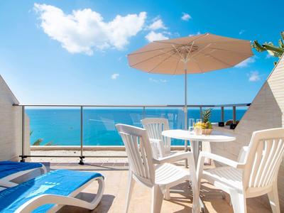 Balkon z białymi krzesłami, stołem, parasolem i widokiem na morze w słoneczny dzień.