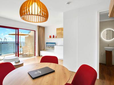 Jasny salon z drewnianym stołem, czerwonymi krzesłami i balkonem z widokiem na morze.