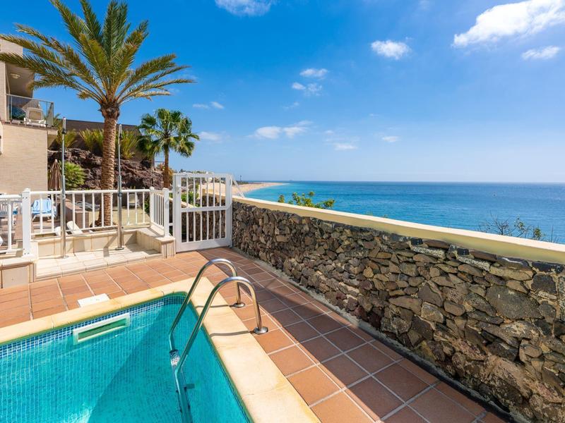 Basen hotelowy z widokiem na morze, taras z stolikami i palmami pod błękitnym niebem.