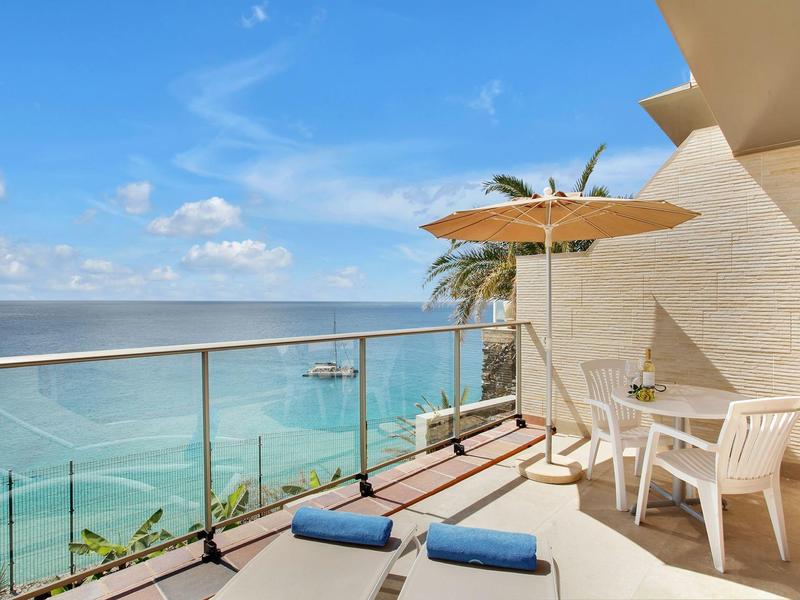 Balkon z białymi krzesłami, parasolem i leżakami z widokiem na morze w słoneczny dzień.