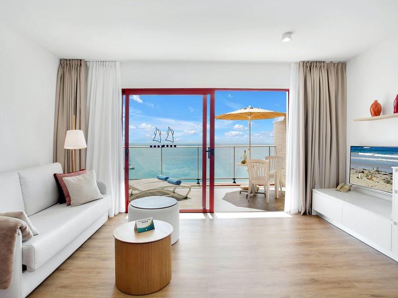 Jasny salon z widokiem na morze, białą sofą, drewnianą podłogą i dostępem do tarasu z parasolem.