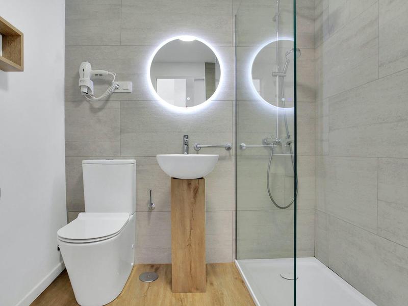 Nowoczesna łazienka z okrągłym podświetlanym lustrem, toaletą i umywalką na drewnianej podstawie.