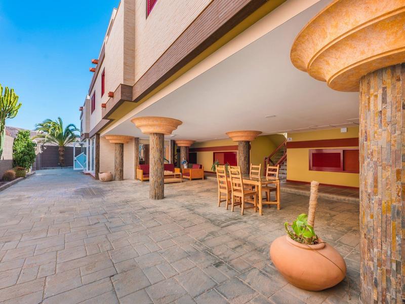 Zadaszony taras z stołem i krzesłami przed hotelem, z rośliną w glinianej doniczce.