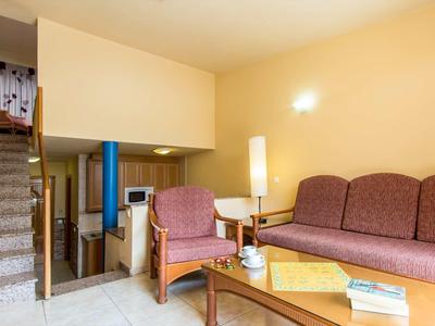 Gemütliches Wohnzimmer mit violettem Sofa, Sessel, Glastisch und einer Treppe im Hintergrund.