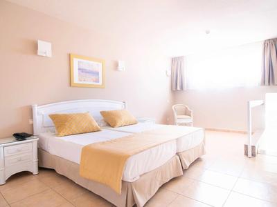 Lichte tweepersoonskamer met wit meubilair, gele accenten en groot raam met gordijnen.