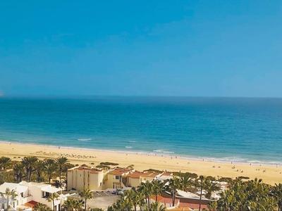Küste mit goldenem Sandstrand, blauem Meer, grünen Bäumen und Gebäuden im Vordergrund.
