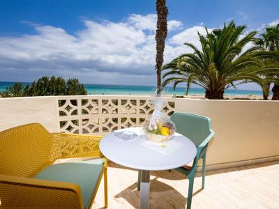 Terrasse mit bunten Stühlen, weißem Tisch und Meerblick, Palmen und bewölktem Himmel.