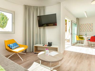 Moderner Wohnraum mit gelbem Sessel, Wandfernseher, kleinem Tisch und Licht durch Fenster.