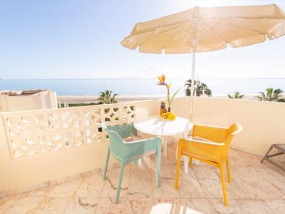 Balkon mit gelbem und türkisfarbenem Stuhl, kleinem Tisch, Sonnenschirm und Meerblick im Hintergrund.