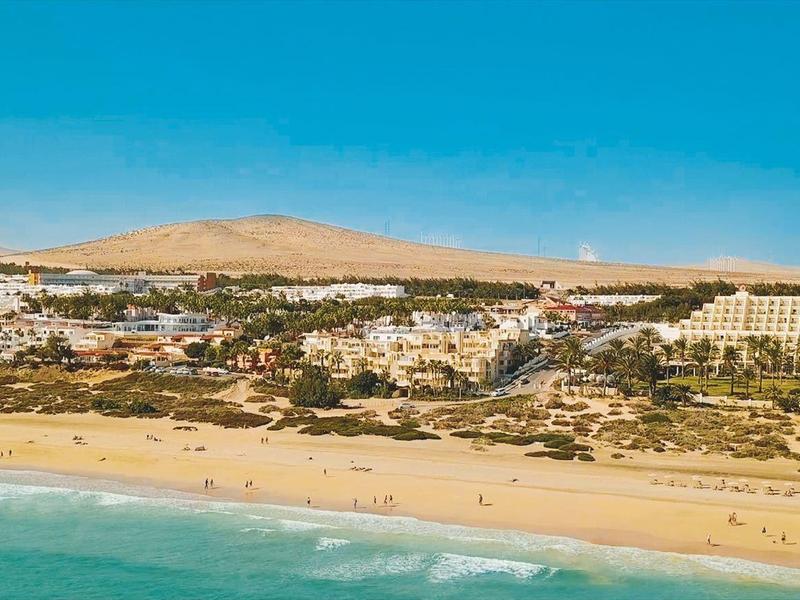 Szeroka piaszczysta plaża z jasnoniebieskim morzem i białymi budynkami ośrodka w tle pod czystym niebem.