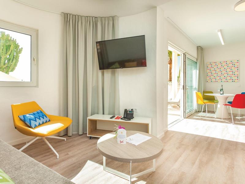 Jasny pokój hotelowy z nowoczesnym krzesłem, telewizorem na ścianie i małym stolikiem na drewnianej podłodze.