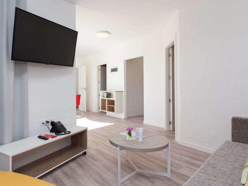 Jasny salon z szarą sofą, okrągłym stolikiem kawowym i telewizorem na ścianie.