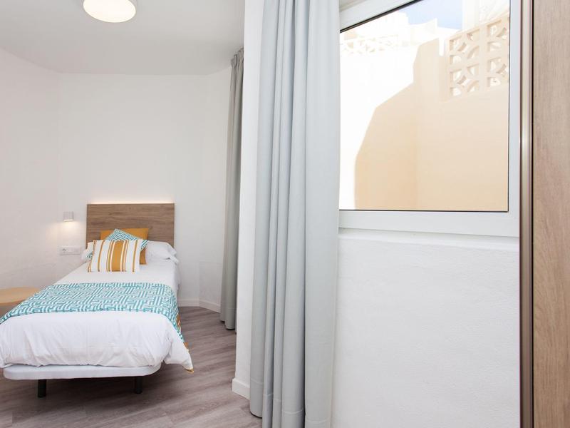 Prosty pokój jednoosobowy z łóżkiem, stolikiem nocnym i oknem obok zasłony.