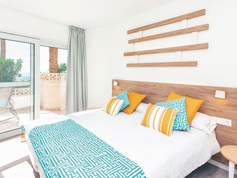 Jasny pokój hotelowy z podwójnym łóżkiem, drewnianym zagłówkiem, kolorowymi poduszkami i balkonem z roślinami.