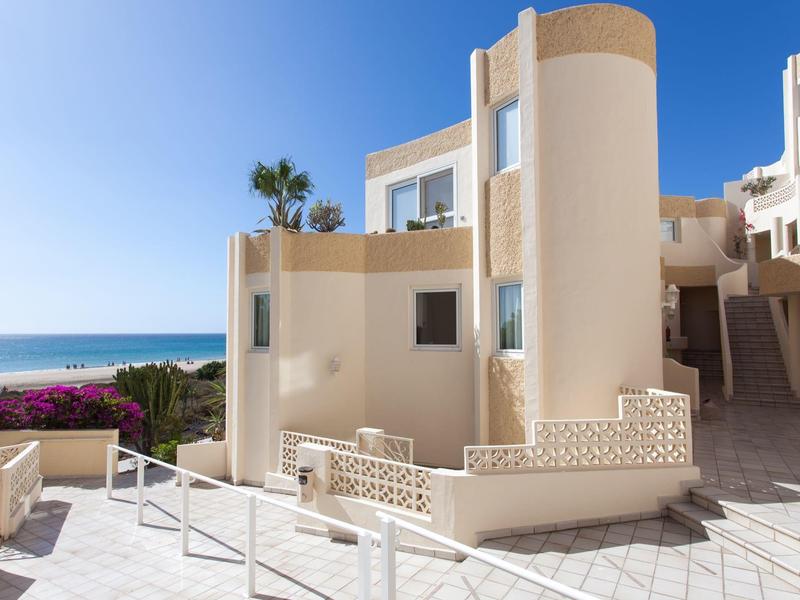 Nowoczesny, piaskowy budynek z okrągłą wieżą i widokiem na morze w słoneczny dzień.