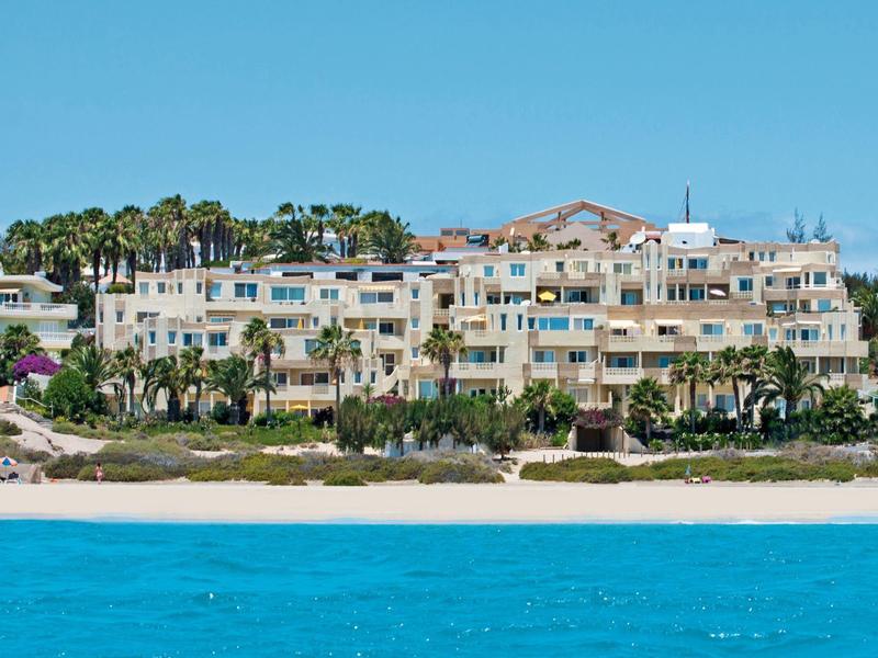 Wielopiętrowy kompleks hotelowy z białymi budynkami i palmami na plaży pod czystym niebem.