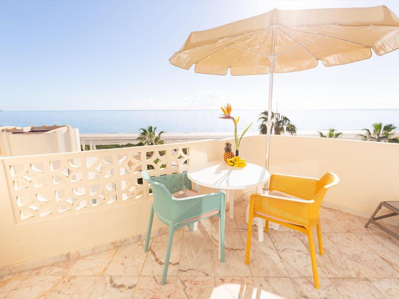 Taras z małym stolikiem, kolorowymi krzesłami i parasolem z widokiem na morze.