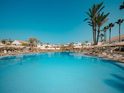 Großer Hotelpool mit Liegestühlen und Palmen unter klarem blauem Himmel.